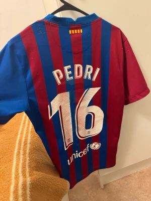 FC Barcelona matchtröja Pedri Nike S - Säljer en officiell FC Barcelona matchtröja från Nike med Pedri och nummer 16 på ryggen. Tröjan har klassiska blå och röda ränder, klubbmärke på bröstet och Rakuten som sponsor. Tillverkad i Dri-FIT material som andas, perfekt för fotboll eller samling.