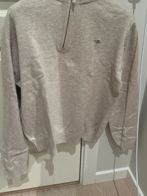 Beige half zip tröja från GANT - Säljer en beige långärmad tröja från GANT med half zip framtill och liten broderad logga på bröstet. Tröjan har ribbade muddar vid hals, ärmslut och nederkant. Perfekt för dig som gillar klassisk och enkel stil. Super laidback och grisch!!