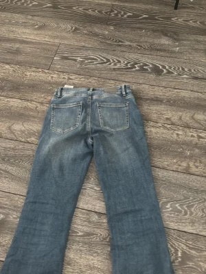 Blå bootcut jeans från Vero Moda - Snygga blå jeans från Vero Moda med klassisk femficksmodell och bootcut-ben. Jeansen har en något högre midja och är tillverkade i mjukt denimtyg. Färgen är mellanblå med lätt slitningar för en cool look.