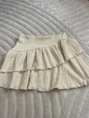 Beige volangkjol från Gina Tricot - Supersöt beige kortkjol från Gina Tricot med dubbla volanger och smock i midjan. Kjolen är gjord i ett strukturerat, lätt material som ger en luftig känsla. Perfekt att styla med en enkel topp eller stickad tröja.