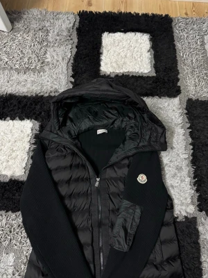 Svart dunväst från Moncler med huva - Säljer en svart dunväst från Moncler med huva och dragkedja framtill. Västen har quiltad design, ribbade partier på sidorna och ett snyggt Moncler-märke på bröstet. Perfekt för lager-på-lager och riktigt clean look.