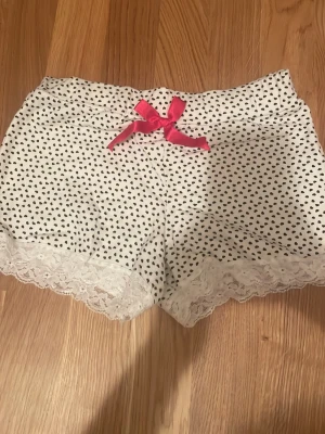 Vita shorts med hjärtan och spets - Supersöta vita shorts med små svarta hjärtan, dekorerade med en rosa rosett i midjan och vit spets längst ner på benen. Perfekta för mysiga dagar hemma eller som pyjamasshorts. säljer pågrund att den aldrig kommit till användning 