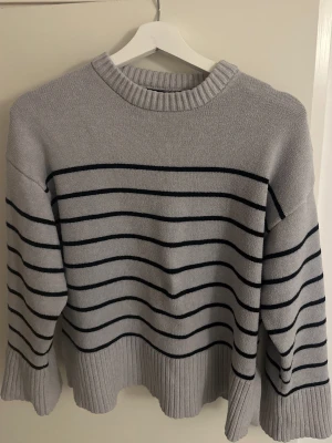 Grå randig stickad tröja - Snygg grå stickad tröja med svarta horisontella ränder. Tröjan har rund hals och breda ribbade muddar vid ärmslut och nederkant. Perfekt för dig som gillar en enkel men stilren look. Passar dig som vill ha en lite loose fit.