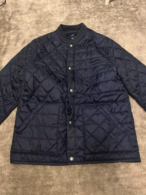 Mörkblå quiltad jacka Polo Ralph Lauren - Säljer en mörkblå quiltad jacka från Polo Ralph Lauren i storlek XL. Jackan har tryckknappar framtill, två stora fickor och ribbade muddar vid ärmslut och krage. Perfekt för kyligare dagar och riktigt snygg till streetwear-stil.