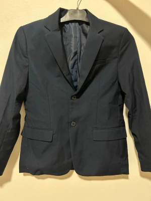 Mörkblå kavaj från H&M, strl 152 - Snygg mörkblå kavaj från H&M i klassisk modell med två knappar framtill, fickor med lock och slits bak. Fodrad insida och stilren passform som passar till många olika looks. Perfekt för dig som vill ha en clean och tidlös stil.