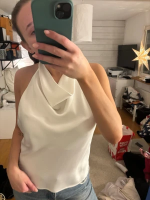Vit halterneck topp från Zara - Säljer en vit halterneck topp från Zara i satinliknande material. Toppen har en snygg draperad front, öppen rygg och knyte i nacken. Perfekt till jeans eller kjol för en trendig look. Stängs med liten hake i ryggen.