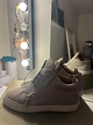 Grå sneakers från Giuseppe Zanotti - Säljer ett par grå sneakers från Giuseppe Zanotti med snygga dragkedjor på sidorna och signaturlogga på plösen. Skorna är i mocka och skinn med vita sulor och rund tå. Perfekta för dig som vill ha en clean men ändå lyxig look.