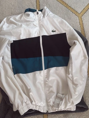 Vit Lacoste jacka med dragkedja och sportig design i kontrastfärger. - Säljer en fräsch vindjacka från Lacoste Sport i vitt med breda block i svart och petrolblått. Jackan har dragkedja framtill, hög krage och elastiska muddar. Klassisk krokodillogo på bröstet och sportig look. Perfekt till vår och höst!
