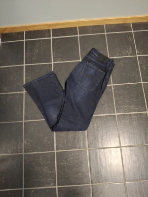Mörkblå bootcut jeans från Nudie Jeans - Snygga mörkblå jeans från Nudie Jeans med klassisk femficksdesign och bootcut-ben. Jeansen har dragkedja och knapp med märkets logga samt subtila slitningar framtill. Perfekta för dig som gillar en tidlös och stilren look.