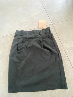 Mörkgrå pennkjol från Kaffe - Snygg mörkgrå pennkjol från Kaffe med resår i midjan och bälteshällor. Kjolen har en stilren look med diskreta fickor bak och är perfekt för dig som gillar enkel men trendig stil. Tillverkad i mjukt material som känns bekvämt hela dagen.