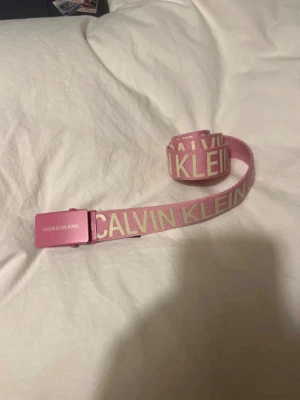 Rosa bälte från Calvin Klein Jeans - Snyggt rosa bälte från Calvin Klein Jeans med tydlig logga i vitt längs hela bandet. Bältet har ett metallspänne i matchande rosa färg och är gjort i slitstarkt syntetmaterial. Perfekt accessoar för att lyfta din outfit.