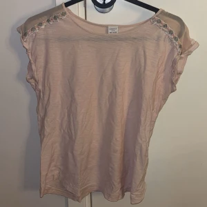 Ljusrosa T-shirt  - Söt ljusrosa T-shirt från Lindex med meshpartier på axlarna och broderade detaljer i färg längs axelsömmen. Från Lindex och storlek 146/152, 10y-12y