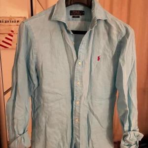 Ljusblå skjorta från Polo Ralph Lauren - Snygg ljusblå skjorta från Polo Ralph Lauren i slim fit-modell. Skjortan har klassisk krage, långa ärmar och knäppning framtill. Ikoniska rosa broderade loggan på bröstet ger en stilren touch. Perfekt för dig som gillar en clean och fräsch look. Nypris cirka 1600kr.