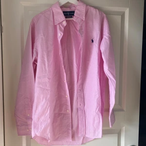 Rosa rutig skjorta från Ralph Lauren - Snygg rosa skjorta från Ralph Lauren med diskret rutigt mönster och klassisk krage. Skjortan har långa ärmar, knappar framtill och den ikoniska blå loggan broderad på bröstet. Perfekt för en clean och stilren look.