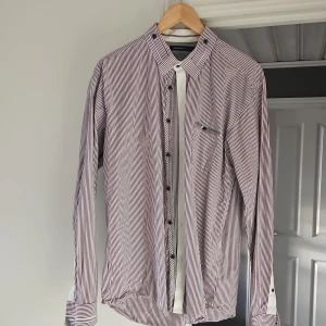 Randig skjorta från Selected Homme - Snygg skjorta från Selected Homme med smala vinröda och vita ränder. Klassisk krage, bröstficka med knapp och svarta knappar framtill. Långärmad modell i mjuk bomull, perfekt för en stilren look.