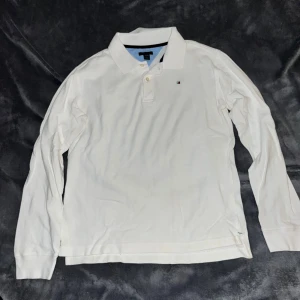 Vit långärmad pikétröja Tommy Hilfiger (BARN) - Vit långärmad pikétröja från Tommy Hilfiger med klassisk krage och knäppning framtill. Liten broderad logga på bröstet. Perfekt för en clean och stilren look. OBS: Barnstorlek, Strl: 16/18.