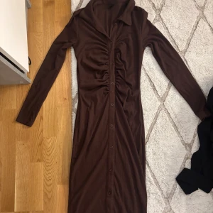 Brun långklänning med knappar - Säljer en brun långklänning med lång ärm och snygg krage. Klänningen har knappar hela vägen framtill och ett rynkat parti längs midjan som ger en cool siluett. Perfekt för dig som gillar stilrena och trendiga plagg.