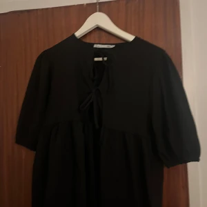 Svart blus med snörning från lager 157 - Svart blus från lager 157 med puffiga korta ärmar och snygg snörning framtill vid halsen. Modellen är luftig och har en avslappnad passform som ger en cool vibe. Perfekt för dig som gillar enkel stil med en twist.