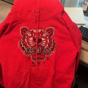 Röd Kenzo hoodie med tigerbrodyr - Säljer en röd hoodie från Kenzo med ikonisk tigerbrodyr och Kenzo Paris-text på bröstet. Tröjan har huva med snörning och en stor magficka. Perfekt statement-plagg för dig som vill sticka ut.