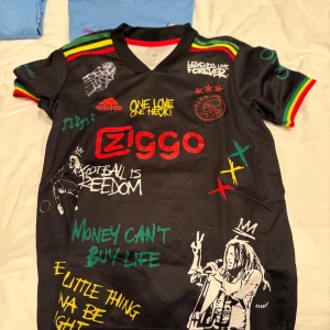 Ajax x Bob Marley svart fotbollströja - Unik svart Ajax fotbollströja från Adidas med färgglada detaljer i rött, gult och grönt. Tryck med Bob Marley, graffiti-texter och Ziggo-logga. Ajax-märke på bröstet och reggaeinspirerade ränder på axlarna. Materialet är lätt och andas, perfekt för match eller streetwear.