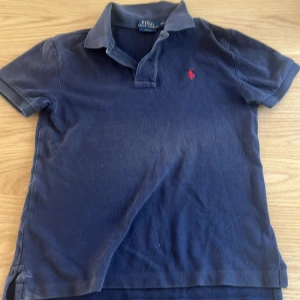 Marinblå pikétröja från Polo Ralph Lauren - Snygg marinblå pikétröja från Polo Ralph Lauren med klassisk krage och korta ärmar. Tröjan har den ikoniska röda logon broderad på bröstet och är gjord i mjuk bomull. Perfekt för en chill och stilren look.