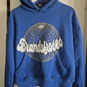 Blå hoodie från Brand of Roses - Snygg blå hoodie från Brand of Roses med stort vitt tryck och globgrafik på bröstet. Boxig och croppad passform, tillverkad i mjuk bomullsfleece för extra komfort. Dubbel fodrad huva och klassisk känguruficka framtill. Perfekt för dig som gillar streetwear.