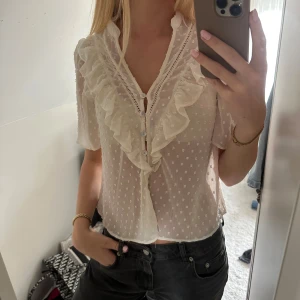 Vit volangblus med prickar från Zara - Supersöt vit blus från Zara med korta ärmar, volangdetaljer längs knäppningen och små prickar i tyget. Blusen är lätt transparent och har knappar framtill. Perfekt för dig som gillar romantisk och feminin stil.