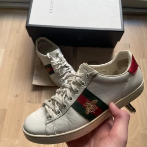 Gucci Ace sneakers med bi-detalj - Gucci Ace sneakers i vitt skinn med klassiska gröna och röda ränder på sidan och broderad guldfärgad bi. Baksidan har metallicröd och metallicgrön häl med ormskinnsmönster och Gucci-logga. Platt sula och snörning framtill. Kommer med originalkartong.