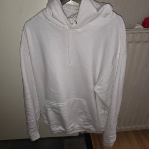 Vit hoodie med svart tryck XL - Vit hoodie i storlek XL med stor svart tryckt design på ryggen. Klassisk känguruficka framtill, snörning i huvan och mjukt material. Perfekt för dig som gillar streetstyle och vill sticka ut med en unik ryggdetalj.