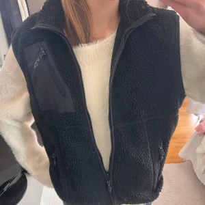 Svart fleeceväst från Noisy May - Mysig svart fleeceväst från Noisy May med hög krage och dragkedja framtill. Västen har flera praktiska fickor med dragkedja, inklusive en sned bröstficka i nylon. Perfekt att slänga över en hoodie eller stickad tröja för extra värme.