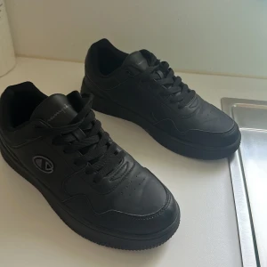 Svarta sneakers från Champion - Säljer ett par helsvarta sneakers från Champion med klassisk design. Skorna har snörning, rund tå och platt sula. Tillverkade i syntetmaterial med diskret Champion-logga på sidan och tungan. Perfekta för en clean och stilren look.