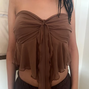 Brun bandeau tubtopp med volang - Säljer en brun bandeau tubtopp med volangkant och knytdetalj framtill. Toppen är ärmlös och har en flowy, luftig passform med lätt transparent tyg. Perfekt för sommardagar och snygg att styla med höga byxor eller kjol. Den har aldrig använts  