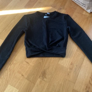 Svart glittrig croppad topp H&M - Svart långärmad kort topp från H&M med glittrigt tyg och twistad detalj framtill. Modellen är croppad och har en rund halsringning. Oanvänd och helt ny