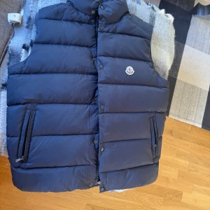Mörkblå tib väst från Moncler - Snygg mörkblå dunväst från Moncler med hög krage, dragkedja och tryckknappar framtill. Västen har två fickor med dragkedja och Moncler-logga på bröstet. Oandvänd. Lappar kvar 