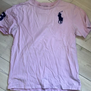 Rosa t-shirt från Polo Ralph Lauren - Snygg rosa t-shirt från Polo Ralph Lauren med klassisk stor marinblå logga på bröstet och tryck på ärmen. Skön passform och tillverkad i mjuk bomull. 