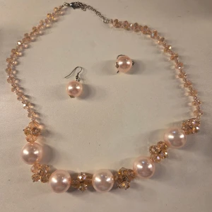Pärlset med halsband och örhängen - Säljer ett superfint smyckeset med halsband och matchande örhängen. Halsbandet har stora ljusrosa pärlor och glittrande kristallpärlor i champagnefärg. Örhängena har stora pärlor i samma ljusrosa ton. Perfekt för dig som vill sticka ut med elegans och lite extra bling.