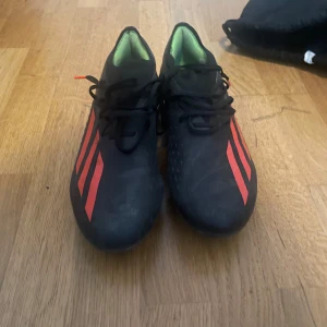 Svarta Adidas fotbollsskor med röda ränder - Säljer ett par svarta Adidas fotbollsskor med klassiska röda ränder på sidorna. Skorna har snörning och är designade för att ge bra grepp på planen. Ovandelen är i syntetmaterial och insidan är ljusgrön. Perfekta för dig som vill sticka ut på matchen.