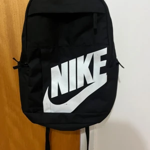 Svart ryggsäck från Nike med stor logga - Snygg svart ryggsäck från Nike med stor vit logga framtill. Väskan har flera fack med dragkedja, vadderade axelremmar och sidofickor. Perfekt för skolan eller gymmet och rymmer allt du behöver till vardagen. Lite färg har gott bort från loggan men, syns inte mycket.