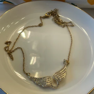 Guldigt halsband med glittriga vingar - Snyggt halsband i guldton med ett hänge format som två vingar täckta av glittrande stenar. Kedjan är tunn och smidig, perfekt för att ge din outfit lite extra bling. Vingarna ger en cool och unik touch som verkligen sticker ut.