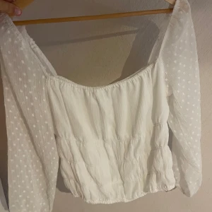 Vit blus med prickiga ärmar Bubbleroom - Supersöt vit blus från Bubbleroom med transparenta, långärmade ärmar i prickigt tyg. Blusen har en rak, något croppad passform och resår vid halsringning och nederkant. Perfekt för dig som gillar romantiska och trendiga detaljer.