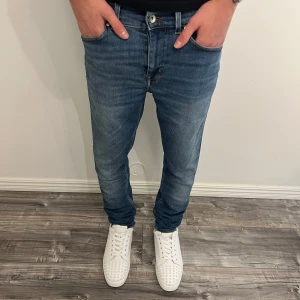 Blå jeans från Tiger of Sweden - Snygga blå jeans från Tiger of Sweden med klassisk femficksdesign och läderpatch bak i midjan. Jeansen har en rak passform med något smalare ben och är tillverkade i mjukt denimtyg med diskreta slitningar för en cool look.