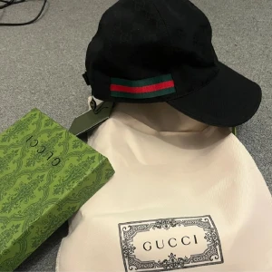 Svart keps med logga gg - Snygg svart keps fmed diskret GG-monogram över hela kepsen och klassisk grön-röd rand på sidan. Kepsen har böjd skärm och justerbar passform. Kommer med originalförpackning och dustbag. Perfekt för dig som vill ha en lyxig streetstyle-accessoar.