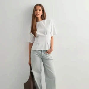 Vit blus med veckad detalj - Säljer en stilren vit blus med korta ärmar och rund hals. Toppen har snygga veckade detaljer framtill som ger en cool och modern look. Passformen är normal och materialet känns mjukt och bekvämt, perfekt till jeans eller kjol.