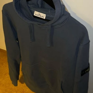 Blå hoodie från Stone Island - Säljer en blå hoodie från Stone Island med klassisk huva och snörning. Tröjan har en stor magficka och den ikoniska loggan på vänster ärm. Perfekt för dig som gillar streetwear och vill ha en stilren look.