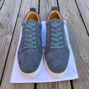 Christian Louboutin grå/blå mockasneakers - Snygga sneakers från Christian Louboutin i grå mocka med klassisk röd sula och vita kanter. Skorna har rund tå, platt sula och snörning. Insidan är beige och de har en stilren, minimalistisk design som passar perfekt till streetwear eller en clean look. Storlek.45