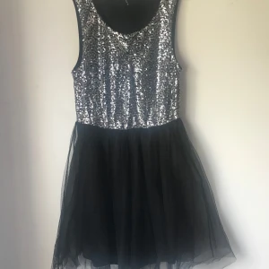 Glittrig svart och silver kortklänning - Supersnygg kort klänning med glittrig silverpaljett-topp och svart tyllkjol. Ärmlös modell med rund halsringning, perfekt för att sticka ut. Kjolen är fluffig och ger en festlig känsla. Ingen dragkedja synlig.