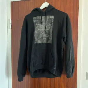 Svart hoodie med tryck och huva - Svart hoodie med stor grafisk print av ett ansikte och texten 'Positivité est priorité' på bröstet. Tröjan har huva med snörning, känguruficka och långa ärmar. Tillverkad i en mix av bomull och polyester, perfekt för en chill och avslappnad stil.