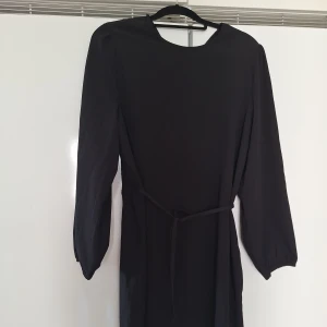 Svart långärmad klänning med knyt i midjan - En stilren svart klänning med långa ärmar och rund hals. Klänningen har ett smalt bälte i midjan som kan knytas för att ge en snygg siluett. Perfekt för dig som gillar enkel och klassisk design. Nyskick. Står XL men passar mer L