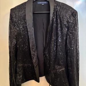 Svart paljettkavaj från French Connection - Glittrig svart kavaj från French Connection med paljetter över hela ytan och klassisk krage i satinliknande material. Kavajen har lång ärm, knäppning framtill och två fickor. Perfekt för att sticka ut med en cool och festlig look.