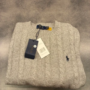 Kapabelstickad polo tröja  - Kabelstickad grå tröja från Polo Ralph Lauren med rund halsringning och klassisk broderad logga på bröstet. Tröjan är i mjuk ullblandning och har ribbade muddar vid ärmslut och nederkant. Perfekt för lager-på-lager under kyliga dagar. Passar nån som e runt 170 